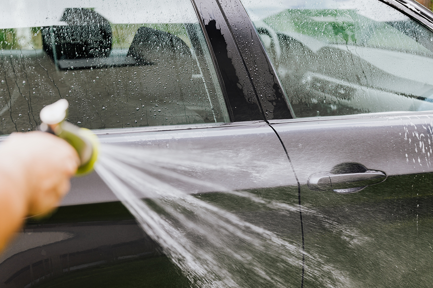 Es posible lavar las ventanillas del coche con vinagre blanco y eliminar pegatinas y adhesivos viejos. El hombre de la foto utiliza una manguera para eliminar el vinagre restante del lateral del coche.